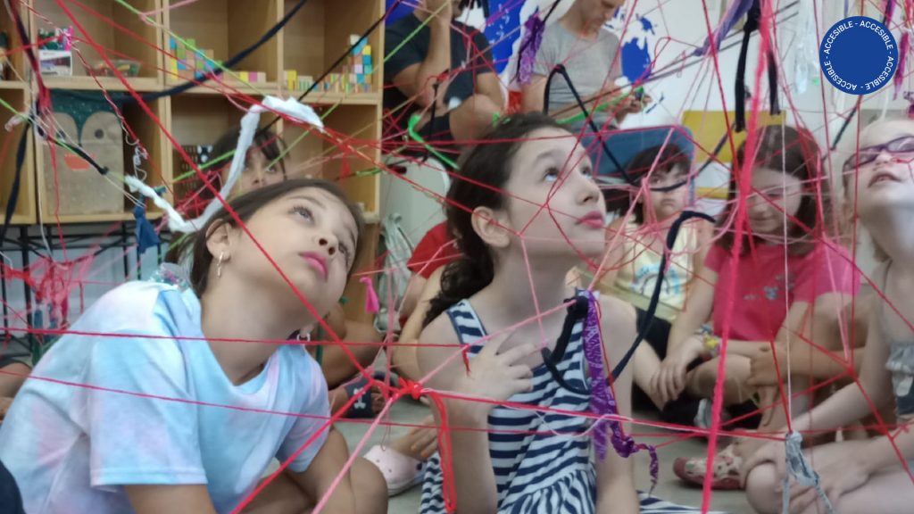 La imagen muestra 3 niñas sentadas en el suelo, frente a una red construida por ellas mismas con hilos de colores, en la sala de educación del museo.